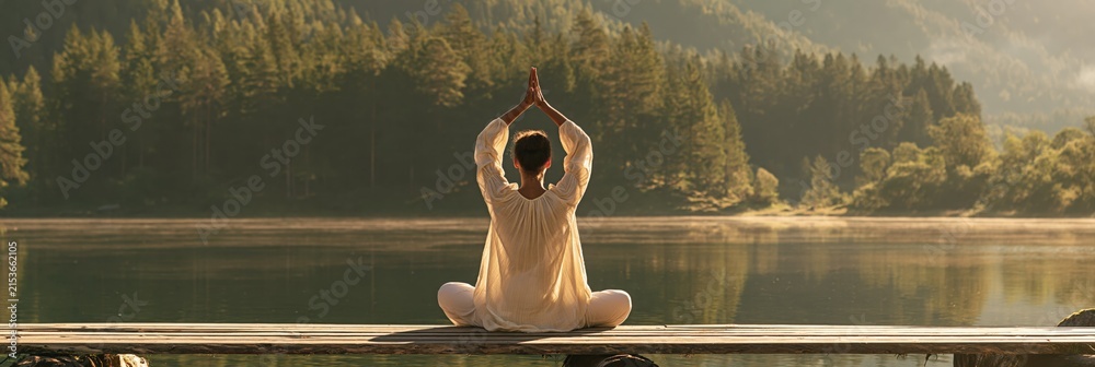 Fototapeta premium Woman seated on a pier doing yoga meditation facing a tranquil lake and forest for mindfulness background and retreat poster