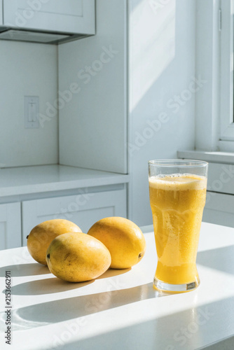 Fresh yellow mangos and glass of mango smoothie on the white table in the white modern kitchen. Copy space