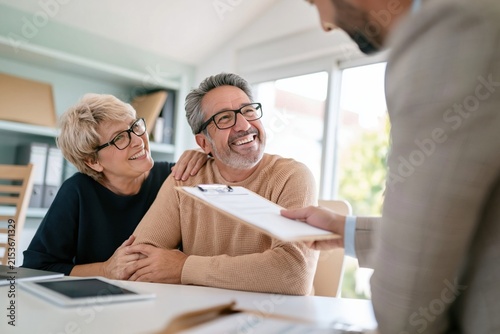 A smiling senior couple sits together at a desk while an adviser presents a clipboard, showing a warm and reassuring atmosphere for financial planning