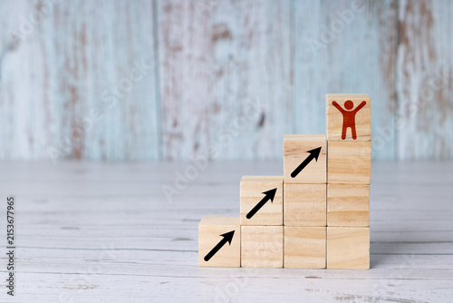 Concept of success, person reaching the top; wooden blocks or cubes arranged like a staircase, with an icon of a person raising their arms in a gesture of success at the top of the stairs.