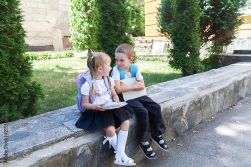 Two happy primary school children with backpacks sitting outdoors near a school building, talking and smiling after class