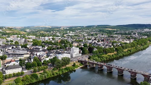 Luftbild von Trier mit der antiken Römerbrücke über die Mosel, umgeben von Stadtarchitektur und grünen Hügeln unter bewölktem Himmel an einem Sommertag.