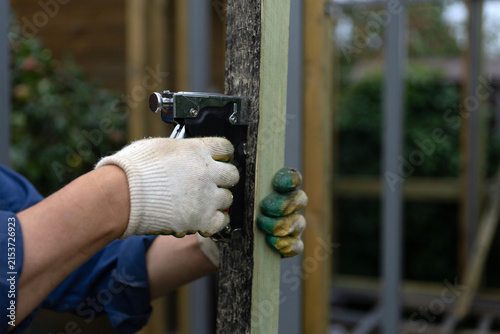A man's hands attach a strip of roofing felt to a wooden beam using a hand-held construction stapler. The wood has been treated with a preservative.