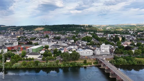 Zeitraffer (Timelapse)  Drohnenaufnahme von Trier mit der antiken Römerbrücke über die Mosel, umgeben von Stadtarchitektur und grünen Hügeln unter bewölktem Himmel an einem Sommertag.