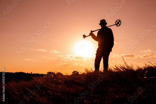 Man Carrying Metal Detector at Sunset, Fiery Orange Background
