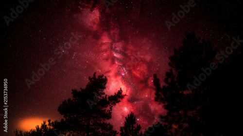 Vibrant red nebula and stars of the Milky Way galaxy glowing above dark forest trees at night, with a subtle orange glow on the horizon, concept of cosmic wonder and vastness
