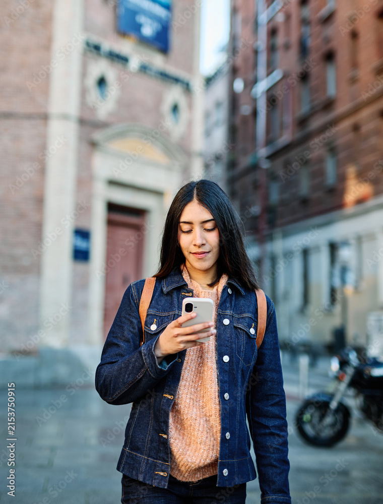 Fototapeta premium A young woman wearing a denim jacket checks her smartphone while standing on a city street. Young woman smiling while holding smartphone and looking up in an urban street