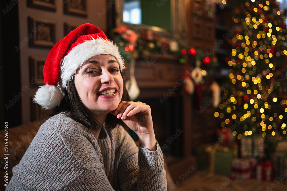 Fototapeta premium Merry Christmas. Young adult woman smiling near Christmas tree in classical dark interior. Happy girl in Santa hat in living room with fireplace Christmas tree gift boxes. Christmas eve at home