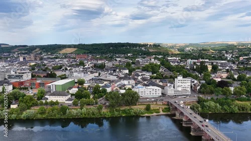 Zeitraffer (Timelapse)  Drohnenaufnahme von Trier mit der antiken Römerbrücke über die Mosel, umgeben von Stadtarchitektur und grünen Hügeln unter bewölktem Himmel an einem Sommertag.