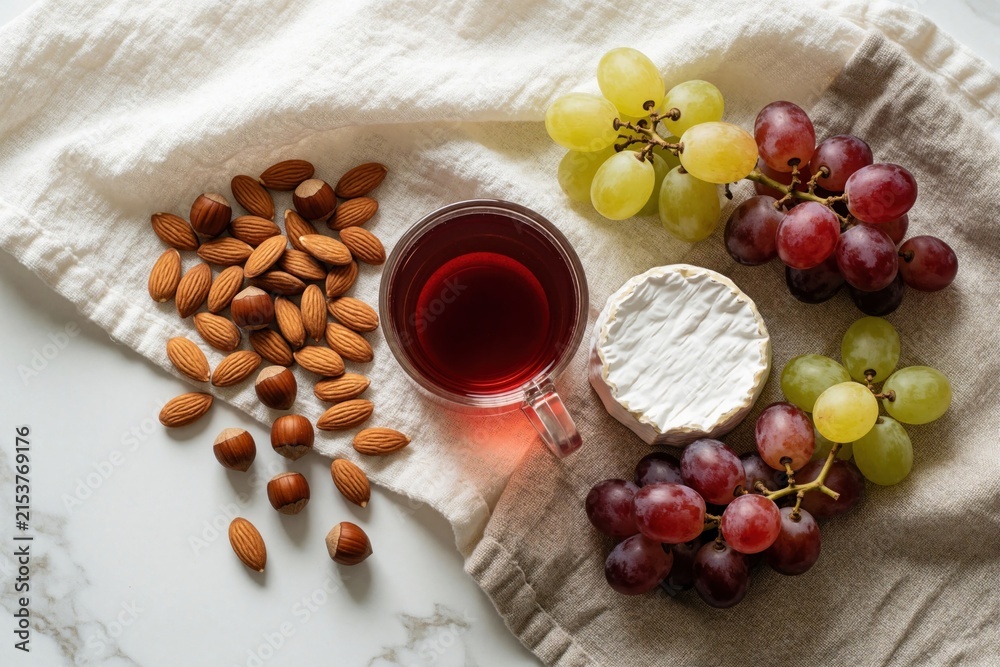 Obraz premium Top view of drink glass with red liquid and snacks. Assortment of fresh grapes, almonds, hazelnuts and brie cheese arranged on textured fabric.