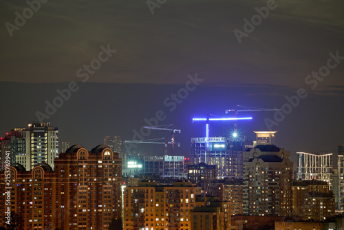 Illuminated city skyline at night with high rise apartment buildings, construction cranes, and modern urban architecture. Real estate development and metropolitan growth concept