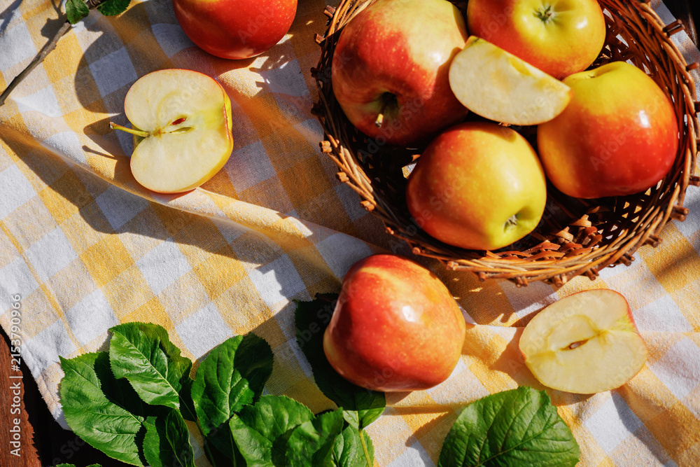 Naklejka premium Fresh red and yellow apples in a wicker basket with sliced apples on a checkered tablecloth in a sunny garden, creating a healthy harvest and autumn lifestyle concept