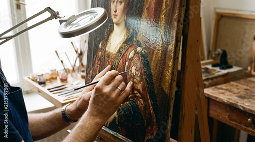 Man art restorer painting on old canvas portrait of woman with brush. Professional restoring ancient oil painting on easel in workshop. Skillful craftsmanship, heritage conservation.