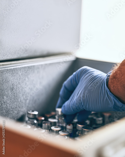Pharmaceutical distribution cold box vaccine vial gloved hand placing temperature logger inside medical cold storage with frost haze, copy space, no face visible