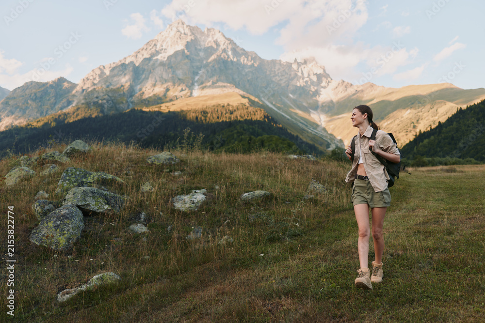 Fototapeta premium Young woman hiking outdoors in casual clothes with backpack walking on grassy trail near rocky mountain under clear sky, active lifestyle and nature exploration
