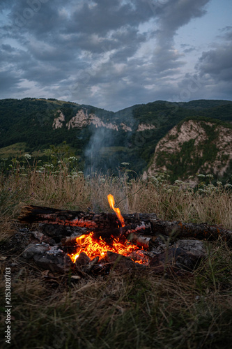 Glowing campfire in a mountain landscape at dusk beneath a dramatic cloudy sky. Perfect for themes of camping, outdoor adventure, hiking, travel, wilderness, survival, and peaceful evenings in nature.
