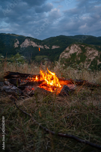 Glowing campfire in a mountain landscape at dusk beneath a dramatic cloudy sky. Perfect for themes of camping, outdoor adventure, hiking, travel, wilderness, survival, and peaceful evenings in nature.