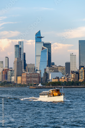 Manhattan II yacht crosses Hudson River on Independence Day. Wooden cabin passenger vessel leaves foamy wake below glass towers and layered evening clouds.