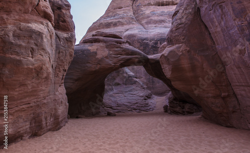 View of the natural sandstone arch at Sand Dune Arch surrounded by towering red rock walls and soft sandy ground in Arches National Park, Grand County, Utah, United States.