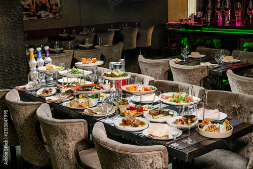 Large table set with many dishes in a restaurant during dinner time. The table is filled with various foods and drinks in a restaurant setting with many empty chairs around.