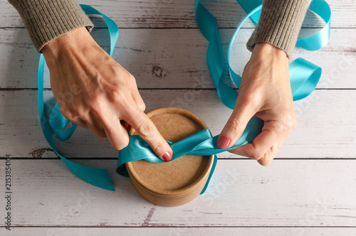 Top view of woman's hands tying blue ribbon on a gift box on white wooden table.