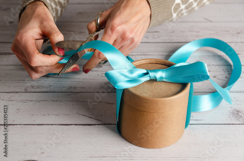 Woman's hands cutting blue ribbon on gift box on a white wooden table.