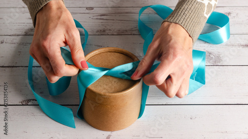 Woman tying blue ribbon on a gift box on a white wooden table.