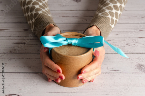 A woman holding a small gift box with blue ribbon on white wooden table.