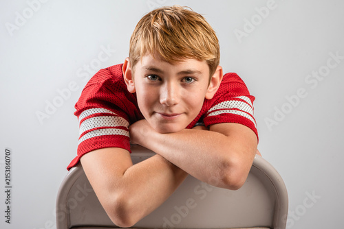 Smiling boy in red American football Jersey leaning over back of a chair with arms crossed