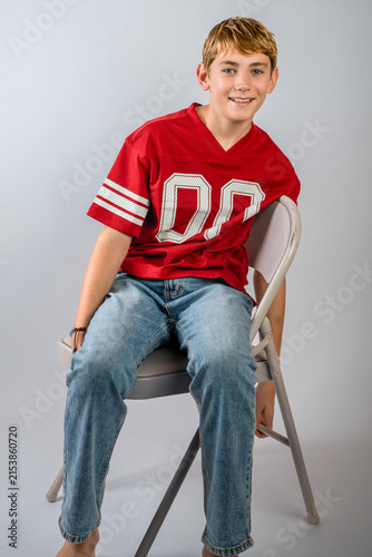 Smiling preteen boy wearing red American football jersey and jeans sitting on folding chair