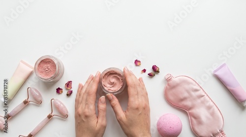 A pair of hands holding a jar of cream, surrounded by skincare products like a face roller, eye mask, and bath bomb, with rose petals scattered around