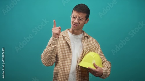 Man wearing glasses and plaid flannel shirt holding yellow hardhat points finger upward with a confident instructive expression in studio; instructional confidence.
