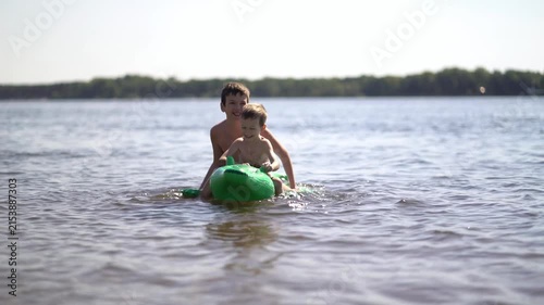 Two young brothers smile and play while floating on a green inflatable crocodile in a calm river on a sunny summer day.