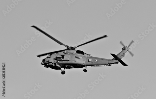 Photography A naval military helicopter flies through a clear blue sky with landing gear lowered during an apparent coastal aviation operation