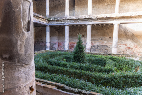 View of the peristyle courtyard with a small evergreen tree in the center of a boxwood hedge maze, surrounded by ancient stone columns and weathered walls in Pompei, Campania, Italy.