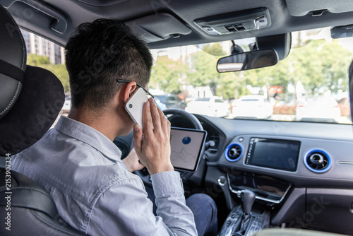 Konstfotografi An adult man in a light shirt sits in a car, holding a mobile phone to his ear, with a tablet on the dashboard