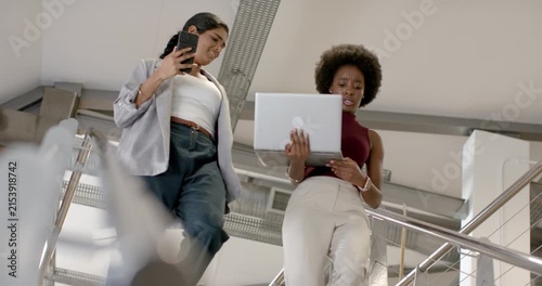 Diverse women coworkers walking down stairwell reviewing brief on laptop and phone in office attire