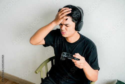 Принти на полотні Stressed Man Wearing Headphones Holding Controller Touches His Forehead in Defea