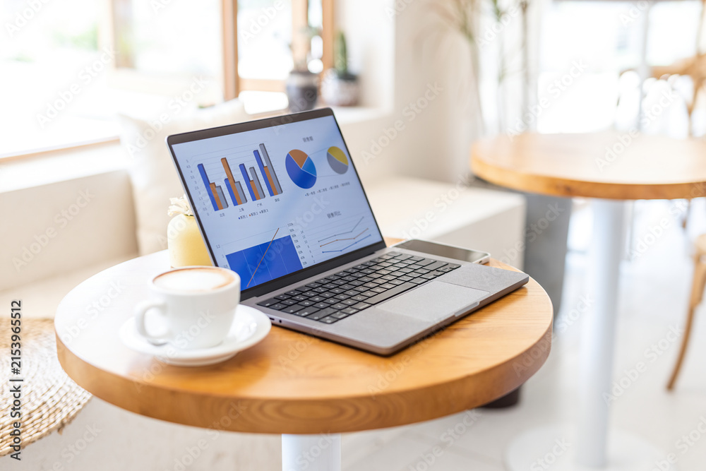 Fototapeta premium A laptop displaying business charts sits on a wooden table next to a coffee cup in a bright cafe workspace.