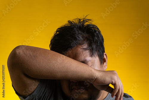 An emotional portrait of a young Asian male hiding his face by burying it into the crook of his folded arm. The subject appears overwhelmed, exhausted, or deeply saddened, positioned