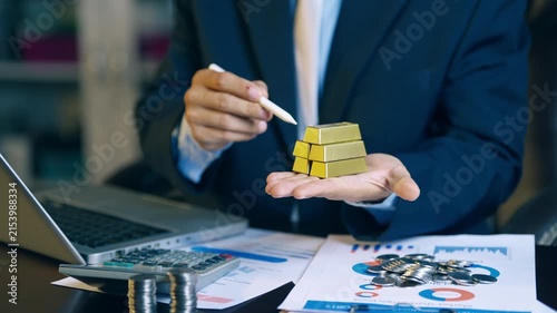Businessman holding and offering gold bars over financial report charts for gold trading and investment concept.
