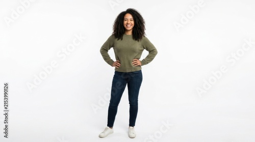 Happy young woman with curly hair standing with hands on hips wearing a casual olive green sweater and blue jeans