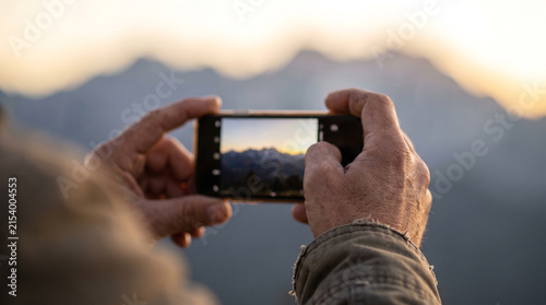 Hands photographing a scenic mountain landscape with a smartphone at sunset, representing travel photography, outdoor adventure and mobile technology use.