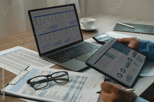 Person uses tablet beside laptop to compare accounting spreadsheets, review printed business documents, check figures with a calculator, and prepare financial statistics, analysis, and reporting.