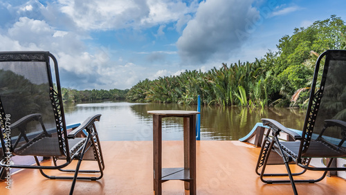 The traditional Indonesian boat klotok floats on the river in the jungle. There are two deck chairs and a table on the deck. There is a riverbed ahead, thickets of nypa fruticans palms along the banks