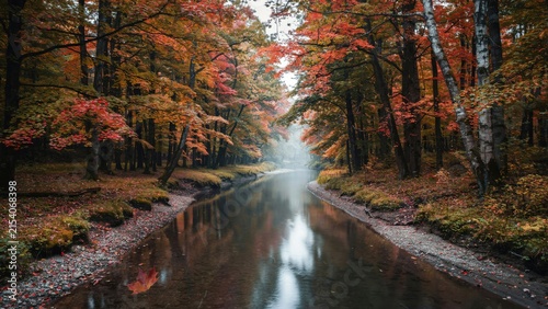 A calm river flowing through a dense forest during autumn with vibrant red and orange leaves on trees and mist hanging over the water surface