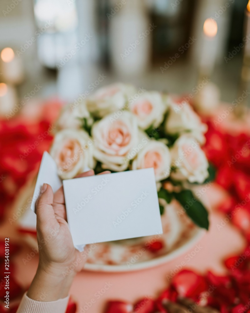 Obraz premium Blank greeting card held in a hand over a table of pink roses for Mothers Day, featuring a mockup for message with a soft romantic atmosphere