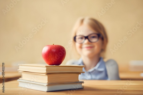 A Young Student Smiling Behind a Stack of Books with a Fresh Red Apple on Top, Signifying Learning and Education in a Classroom Environment