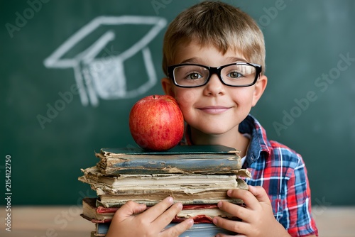 A Young Boy with Glasses Smiles While Holding a Stack of Old Books and an Apple in Front of a Chalkboard with a Graduation Cap Drawing