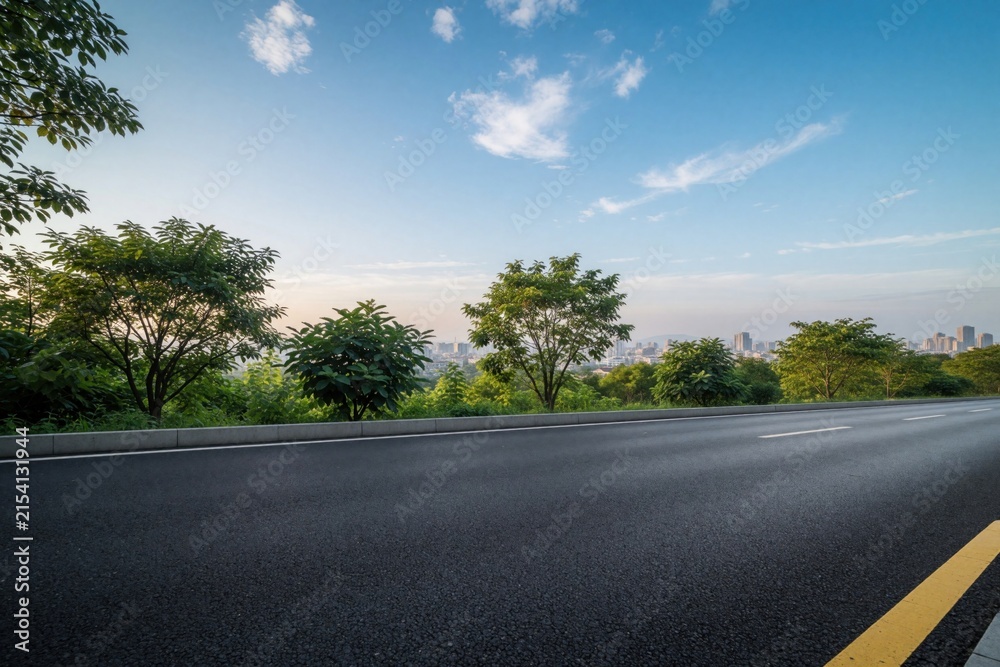 Fototapeta premium Empty asphalt highway with white lane markings stretches toward urban skyline. Modern city buildings, skyscrapers appear under blue sky with clouds. Green trees line roadside near empty street in